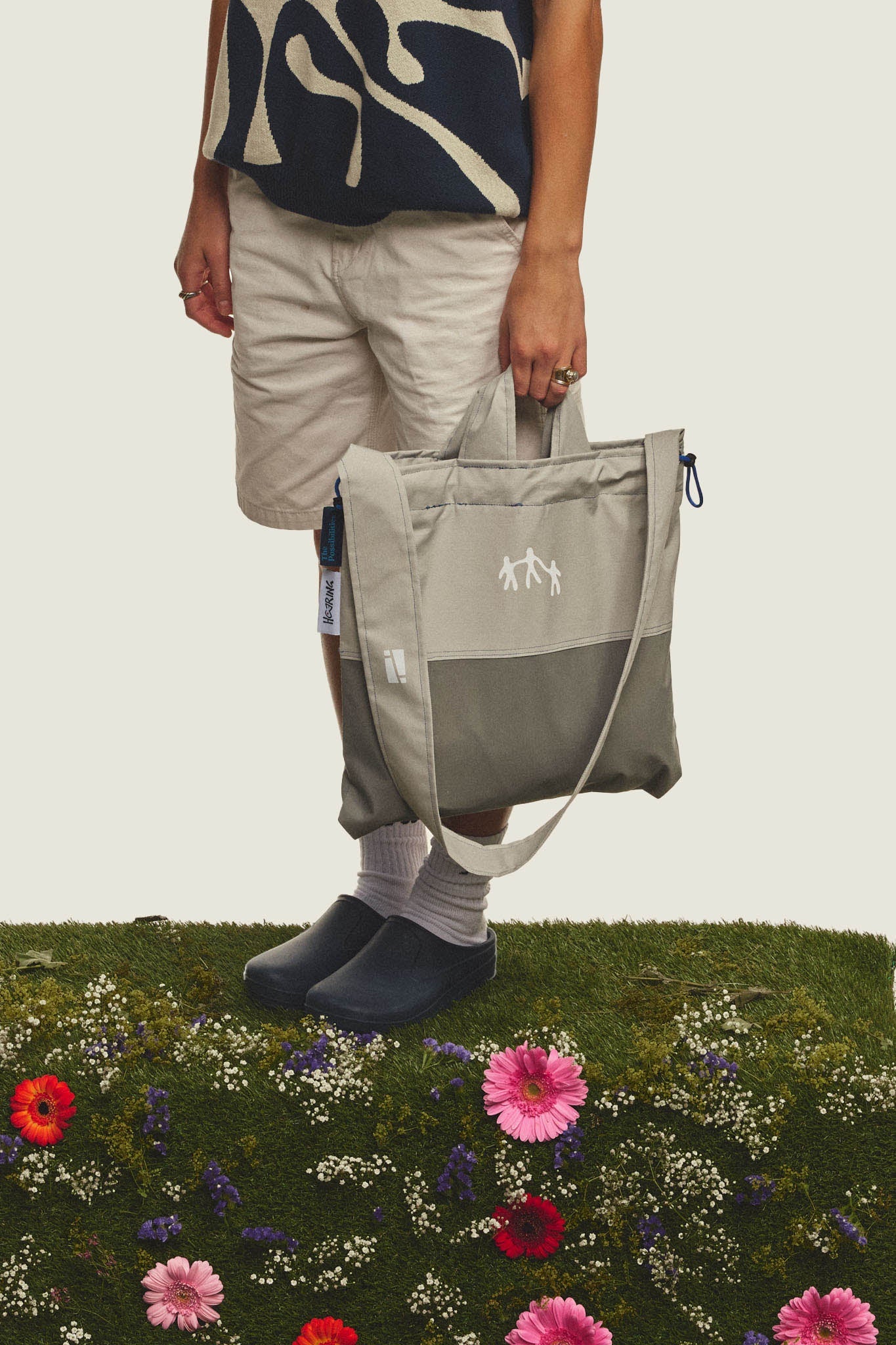 Editorial campaign photo of a person holding a sustainable tote bag, from the collab collection between Studio Haering and The Possibilities Clothing, in a green meadow full of flowers.